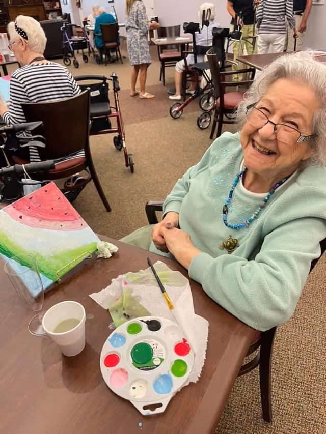 An elderly woman with gray hair and glasses smiling while sitting at a table with painting supplies, including a paint palette, brush, and a partially painted canvas. Other elderly people are visible in the background, some seated and some standing, in a communal room with chairs and tables.