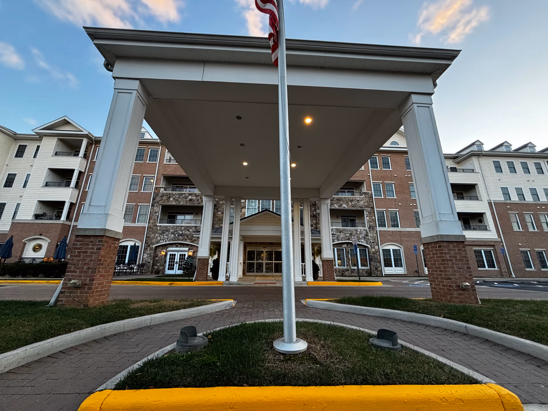 Front entrance of a multi-story senior living facility with a covered drop-off area supported by white columns and brick bases, an American flagpole in the center, and a building facade featuring brick and stone accents under a partly cloudy sky.