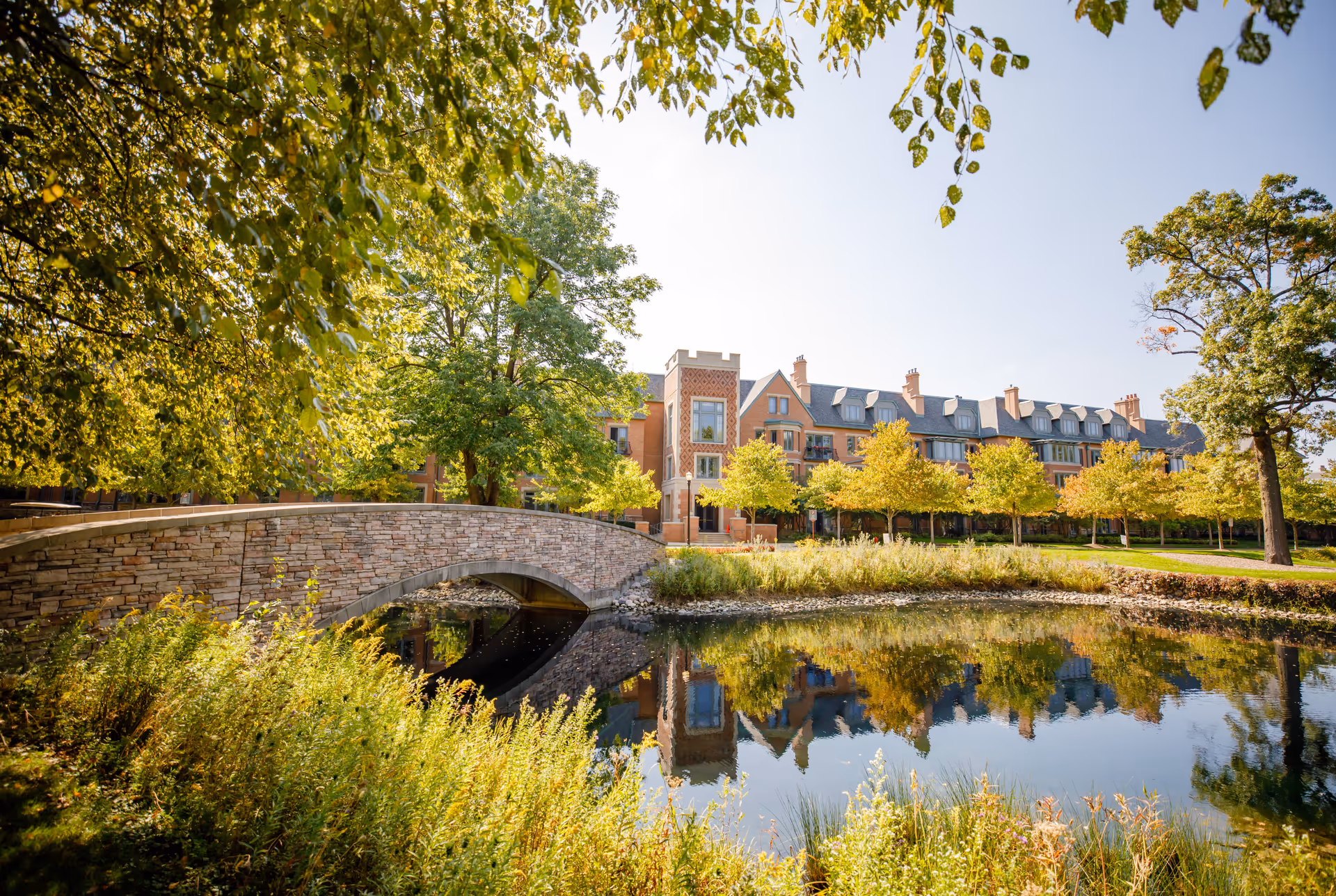 A scenic outdoor view of The Garlands of Barrington featuring a stone arch bridge over a calm pond with reflections of trees and a large brick building in the background under a clear sky.