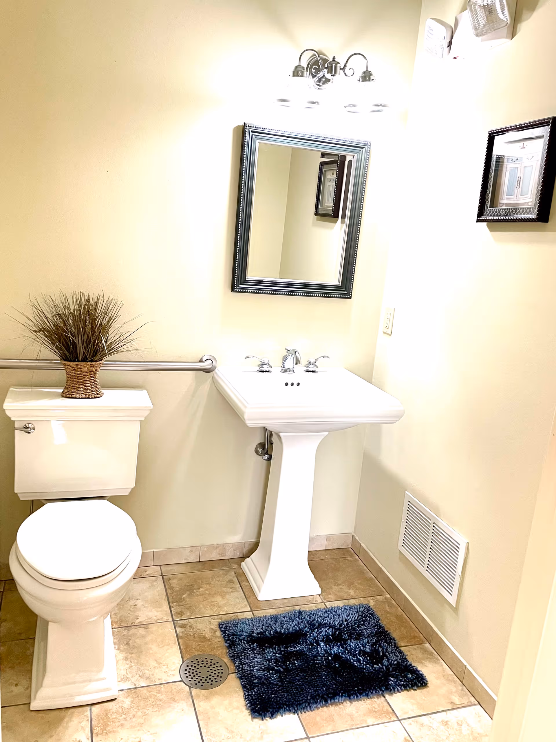 A clean bathroom with beige tiled floor and cream-colored walls. It features a white toilet with a small plant in a wicker basket on top, a white pedestal sink with chrome faucets, a rectangular mirror with a dark frame above the sink, a blue textured bath mat on the floor, a metal grab bar on the wall behind the toilet, and a wall vent near the floor.