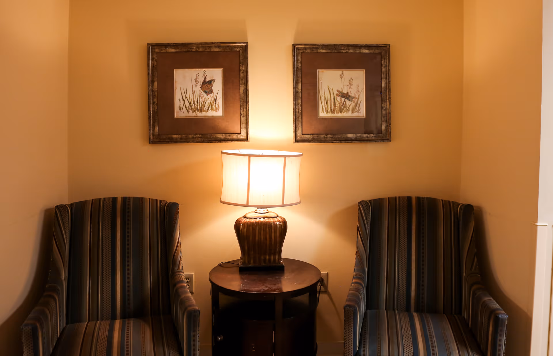 Two striped armchairs flank a small round table with a lit lamp beneath two framed botanical prints on a beige wall.
