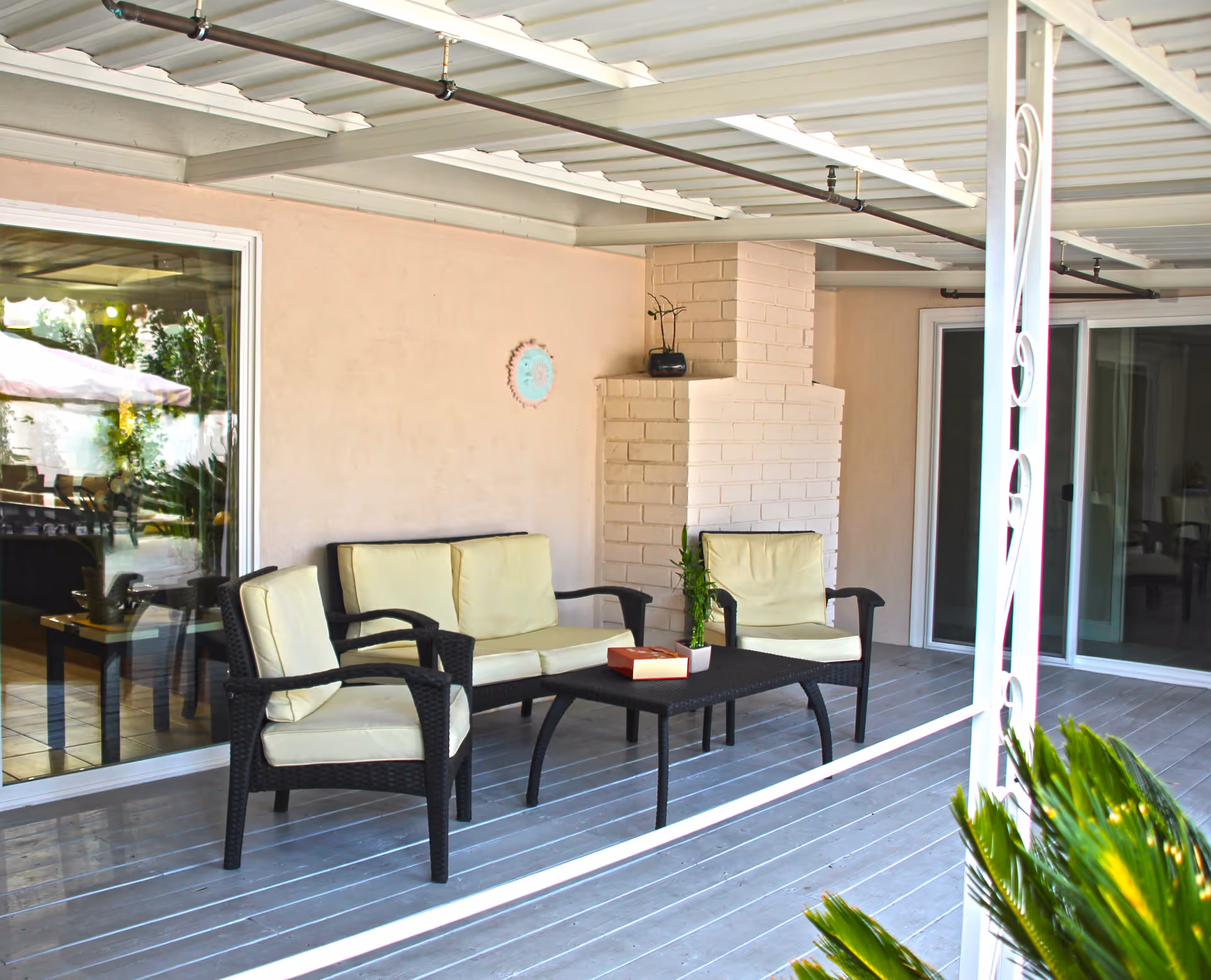 Covered outdoor patio area with a seating arrangement consisting of two armchairs and a loveseat with light-colored cushions around a black coffee table. A small potted plant and a box are on the table. The patio has a light gray wooden floor, a white brick chimney, and sliding glass doors leading inside. Some greenery is visible in the foreground and through the glass doors.