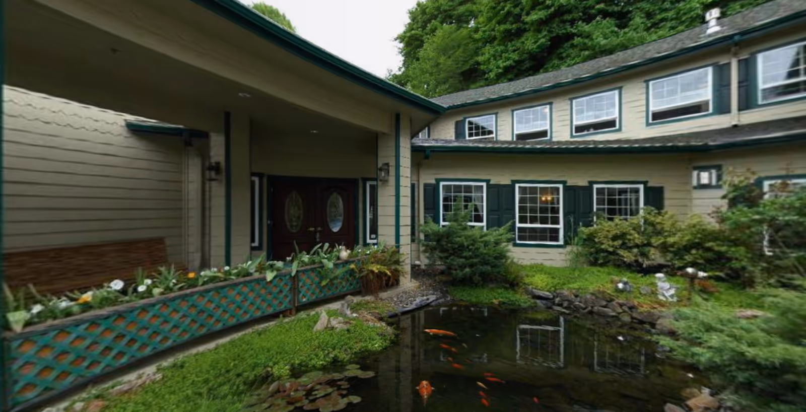 Exterior view of The Amber facility showing a beige building with green trim, multiple windows, a covered entrance with double doors, a small pond with koi fish, and surrounding greenery including bushes and trees.