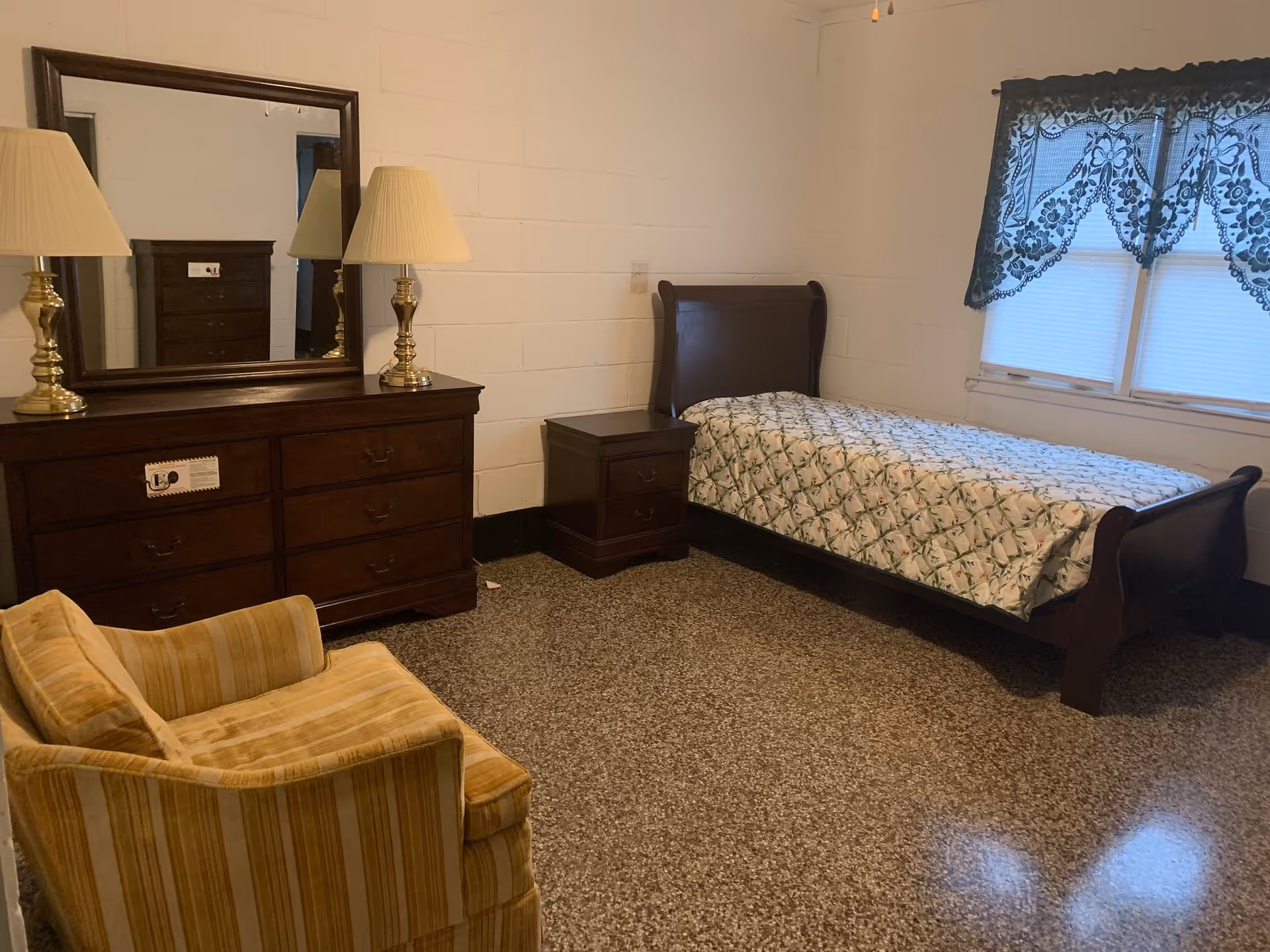 A senior living facility bedroom with a single wooden bed covered with a floral bedspread, a wooden nightstand, a wooden dresser with a large mirror and two lamps, and a yellow striped armchair. The room has white painted brick walls, a window with blue lace curtains, and a speckled floor.