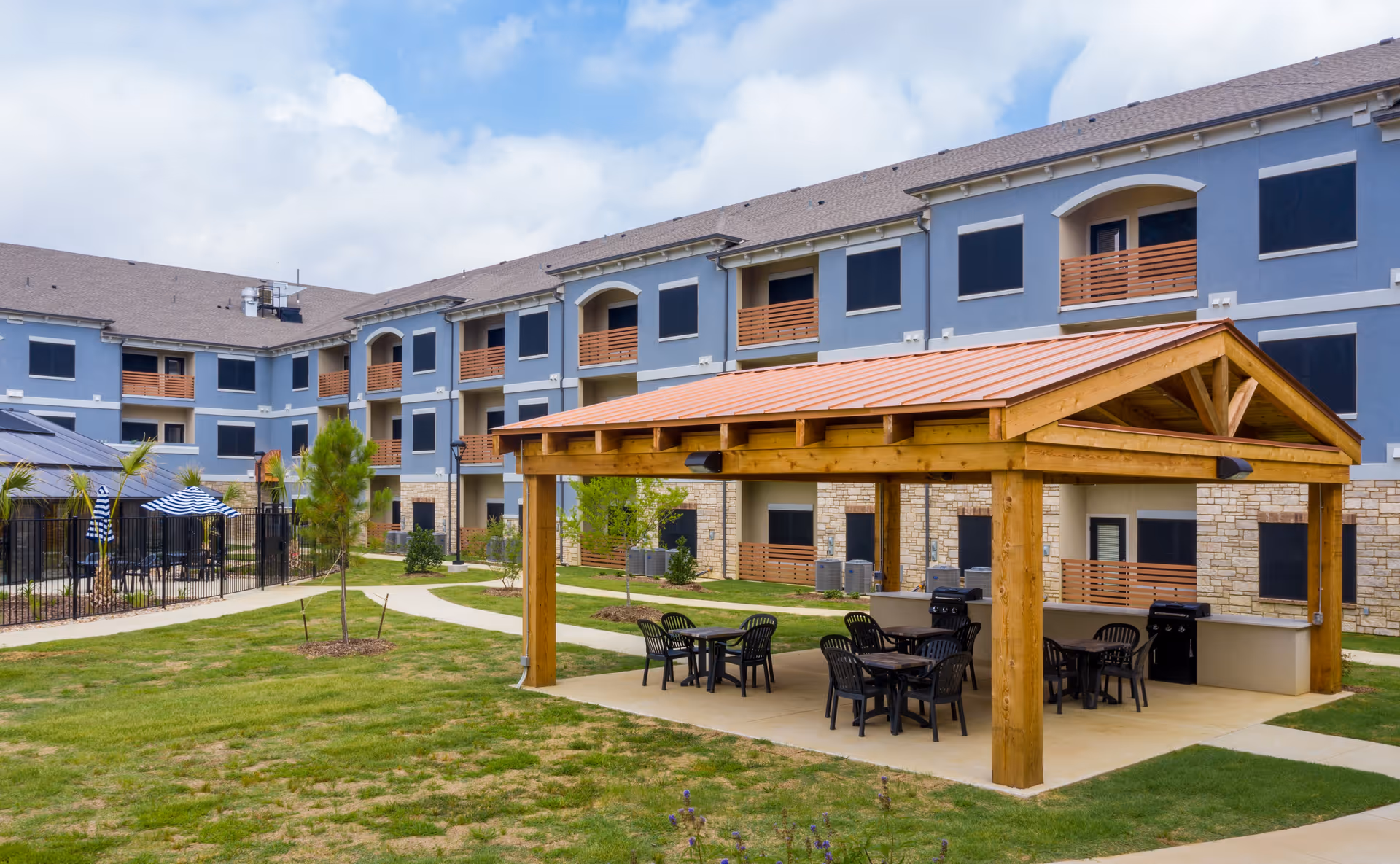 Outdoor courtyard of a senior living facility with a wooden pavilion, tables and chairs, and a three-story building with balconies in the background.