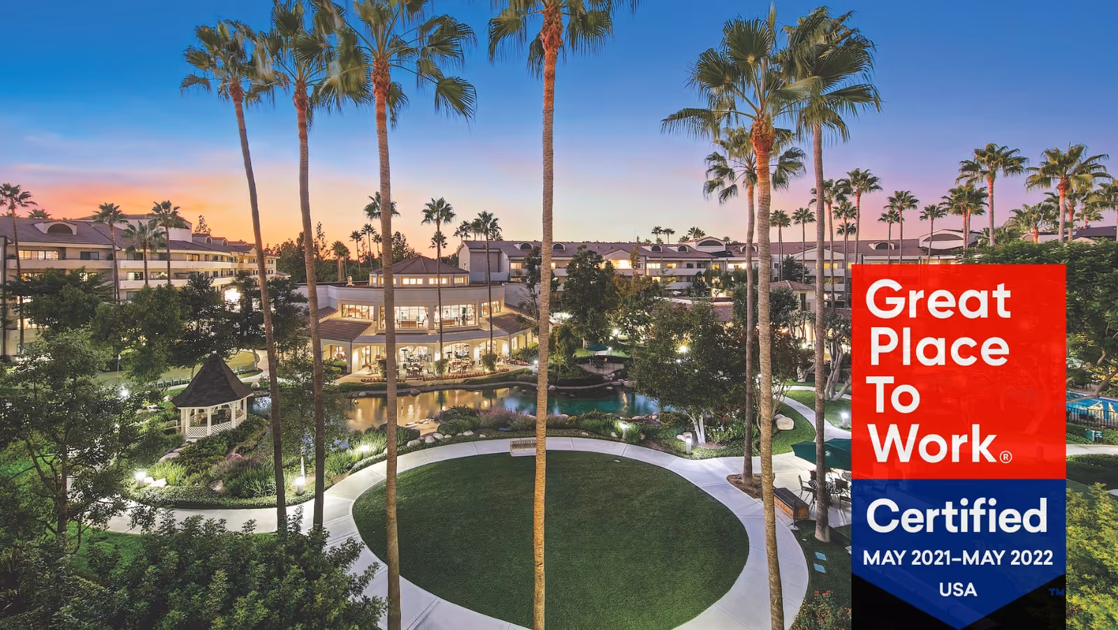 Aerial view of Morningside of Fullerton senior living facility at sunset, showing a landscaped garden with tall palm trees, a gazebo, walking paths, a pond, and multi-story buildings in the background.