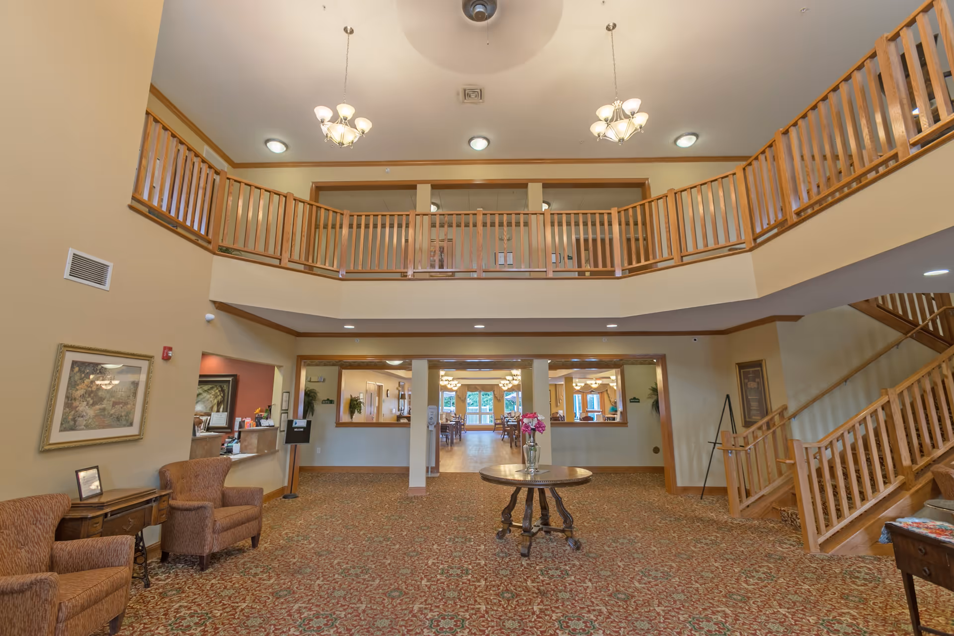 Spacious lobby area of a senior living facility with a high ceiling and wooden balcony railing on the upper level. The room features patterned carpet, two armchairs with a small table on the left, a round table with a vase of flowers in the center, and a staircase with wooden railings on the right. The back wall has large windows and an open doorway leading to a dining area with tables and chairs.