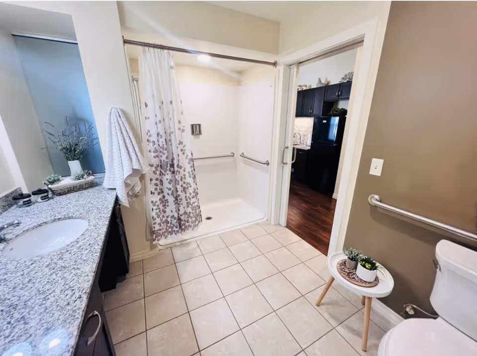 A clean and accessible bathroom featuring a walk-in shower with a floral shower curtain, grab bars, a granite countertop with a sink, a towel hanging on the wall, a toilet with a grab bar beside it, and a small round table with potted plants. The bathroom opens into a kitchen area with dark cabinets and a black refrigerator.