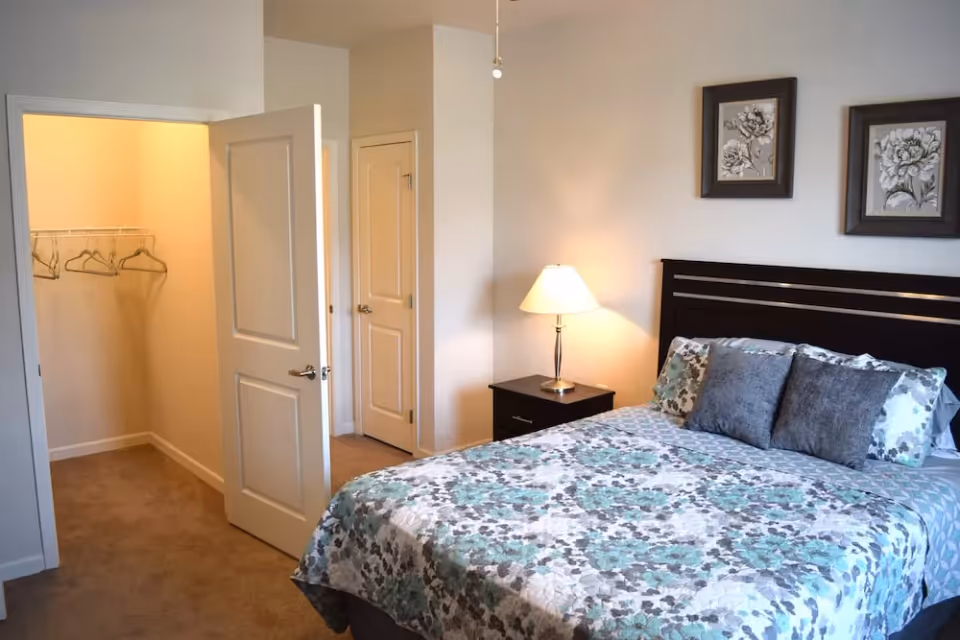 A bedroom with a bed covered in a floral patterned bedspread in shades of blue, gray, and white. There are two gray pillows and two floral pillows on the bed. A nightstand with a lamp is next to the bed. The room has beige walls and carpet. Two framed floral artworks hang above the bed. An open door reveals a walk-in closet with hangers inside.