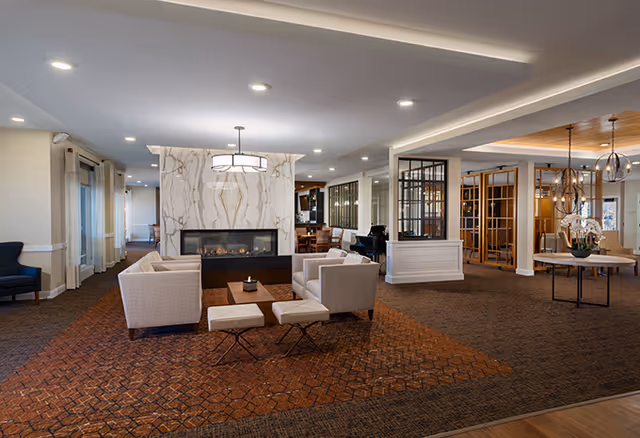A spacious senior living facility common area with a modern gas fireplace surrounded by a marble wall. The room features beige armchairs and ottomans arranged around a small wooden coffee table on a patterned carpet. To the right, there is a round table with a flower arrangement and decorative pendant lights hanging from the ceiling. The space is well-lit with recessed lighting and has large windows with curtains along the left side.