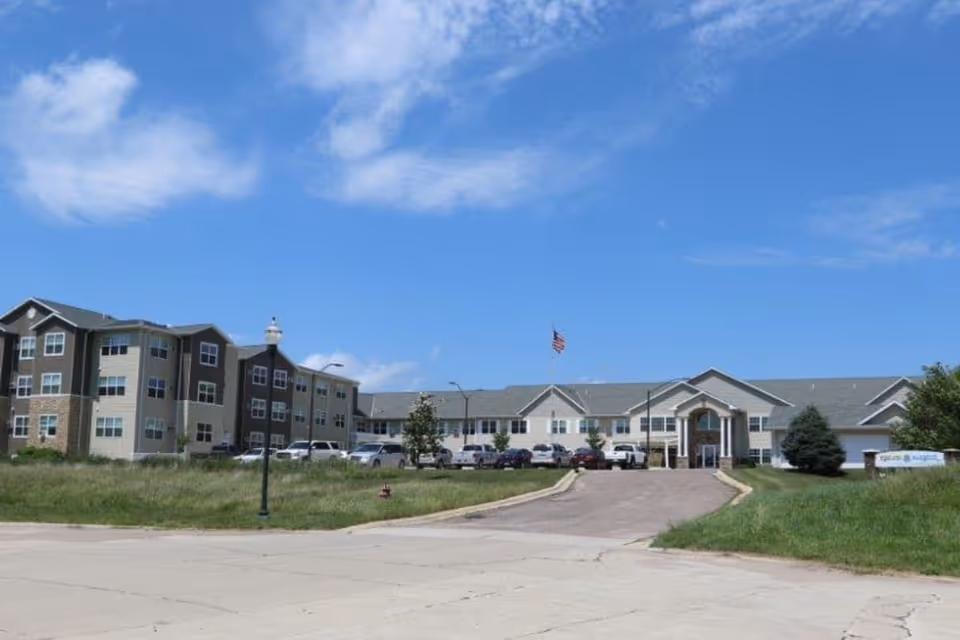 Exterior view of a large senior living facility named Keelson Harbour under a blue sky with scattered clouds. The building is multi-story with beige and brown siding, multiple windows, and a central entrance with a covered portico. Several cars are parked in front of the building, and an American flag flies on a flagpole near the entrance.