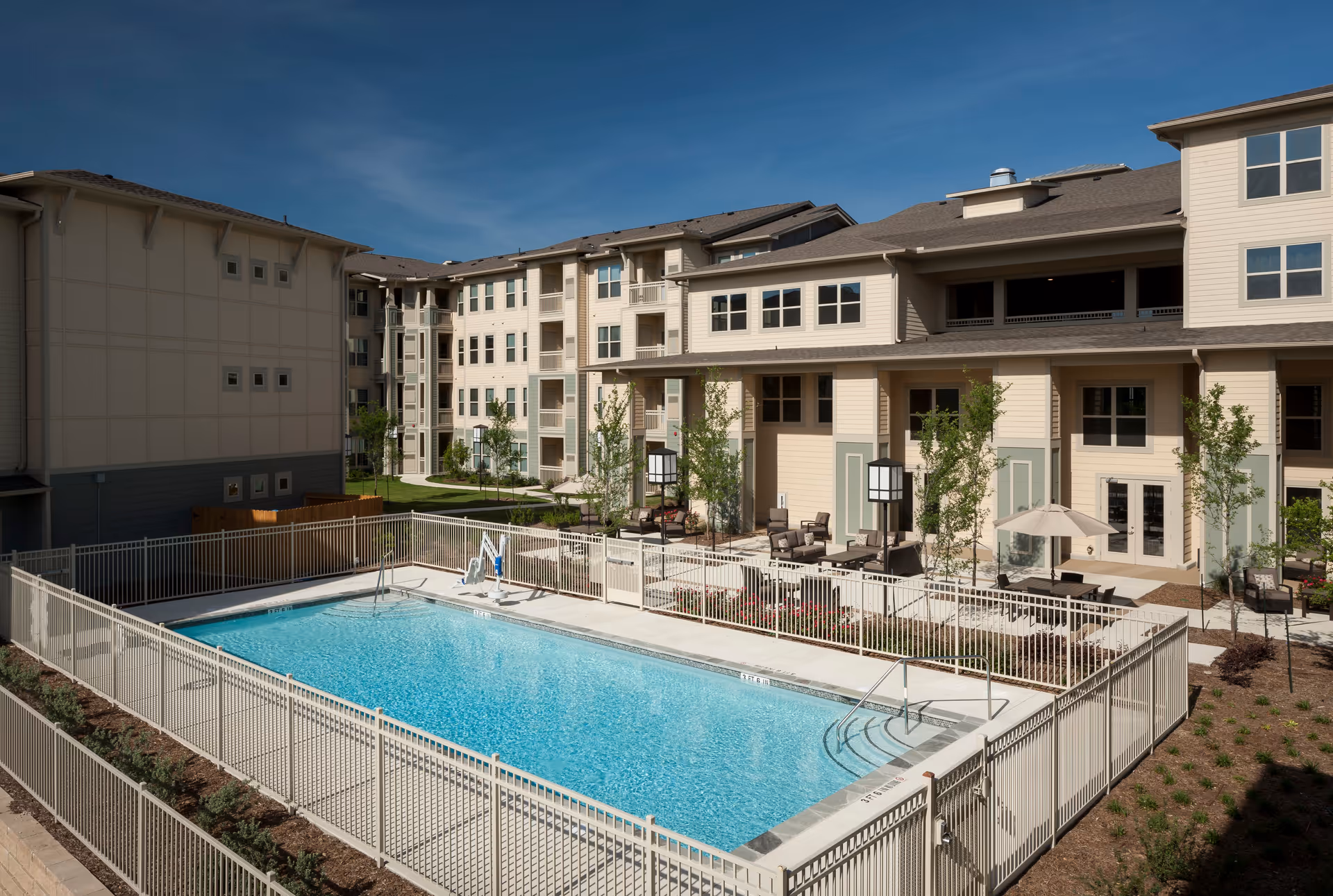 Outdoor swimming pool area enclosed by a white metal fence with lounge chairs, tables with umbrellas, and a multi-story residential building in the background under a clear blue sky.