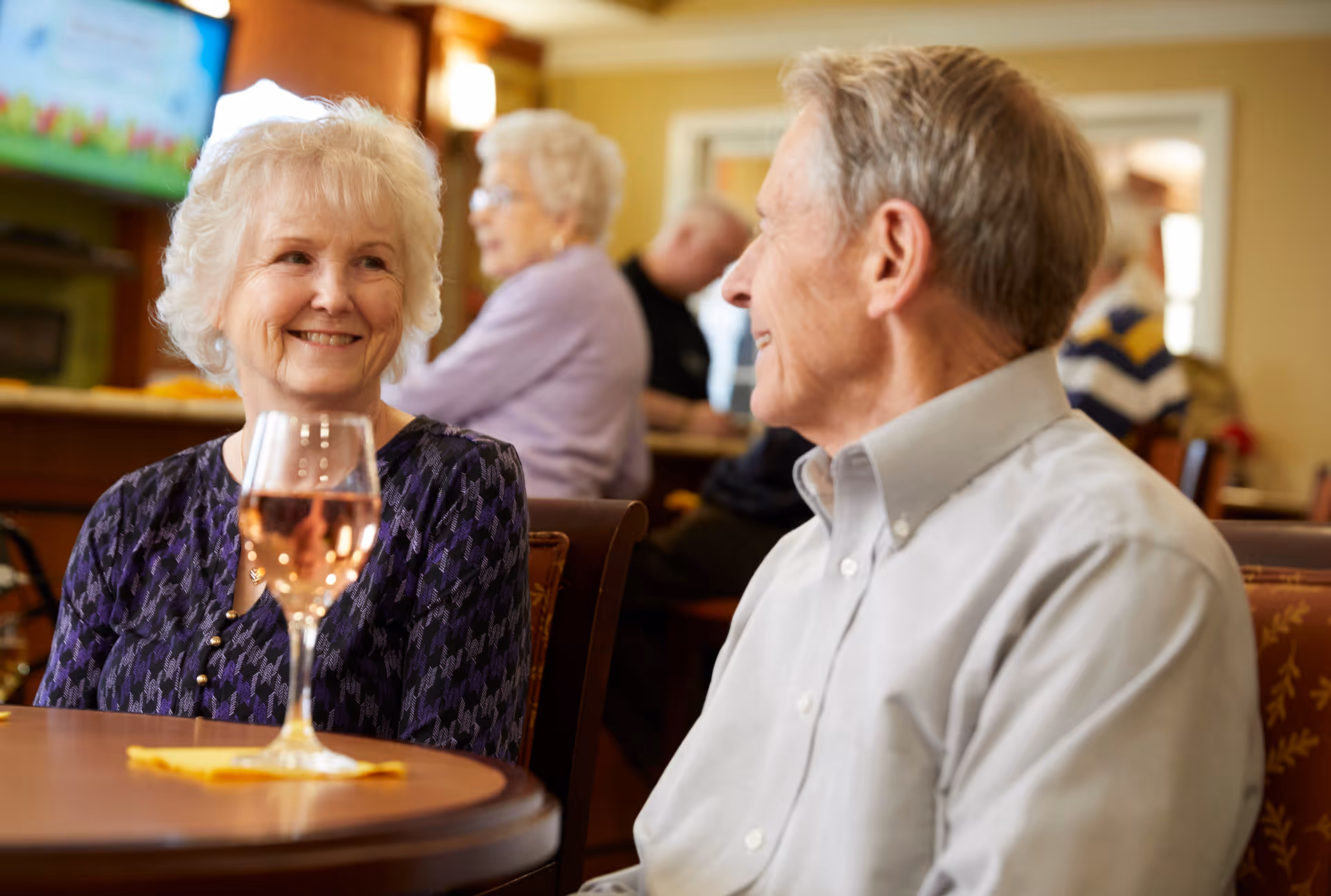 An elderly woman and man sitting at a table in a senior living facility dining area, smiling and engaging in conversation. The woman has a glass of rosé wine in front of her. Other seniors are visible in the background, seated and interacting in a warmly lit room.