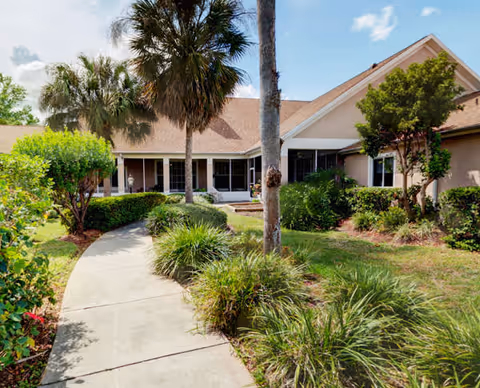 Front entrance of a single-story assisted living building with a curved concrete walkway, palm trees, and landscaped greenery.