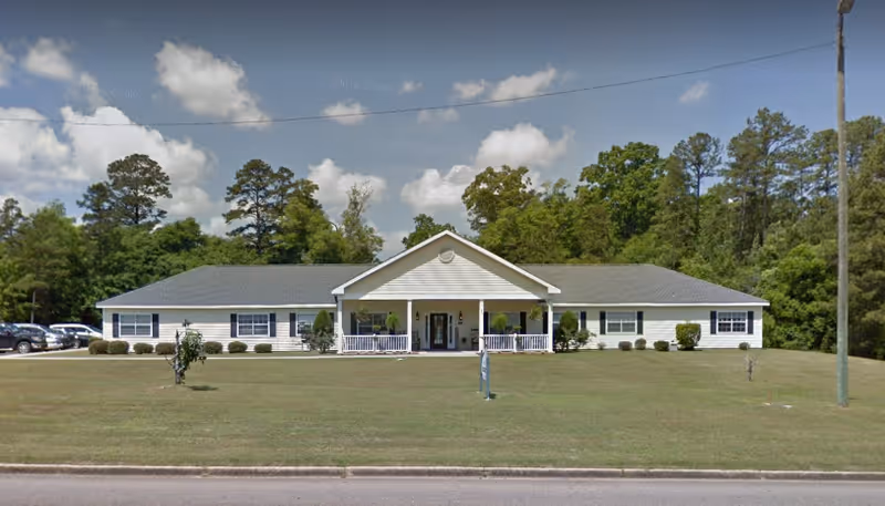 Single-story white building with a gray roof and a covered front porch, surrounded by a large grassy lawn and trees in the background under a partly cloudy sky.