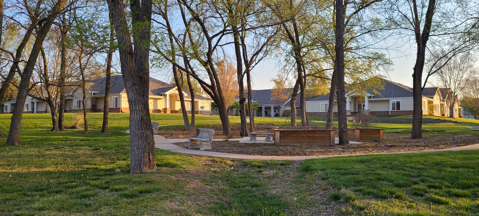 Outdoor view of a senior living facility named Maria Court, showing a grassy area with trees, a paved walking path, stone benches, and raised garden beds. The facility buildings are visible in the background under a clear sky.