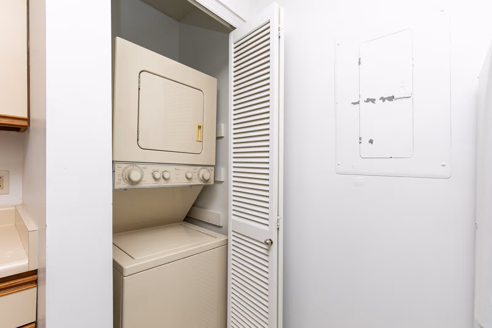 A small laundry area with a stacked beige washer and dryer unit inside a closet with a white louvered door, adjacent to a white wall with an electrical panel.