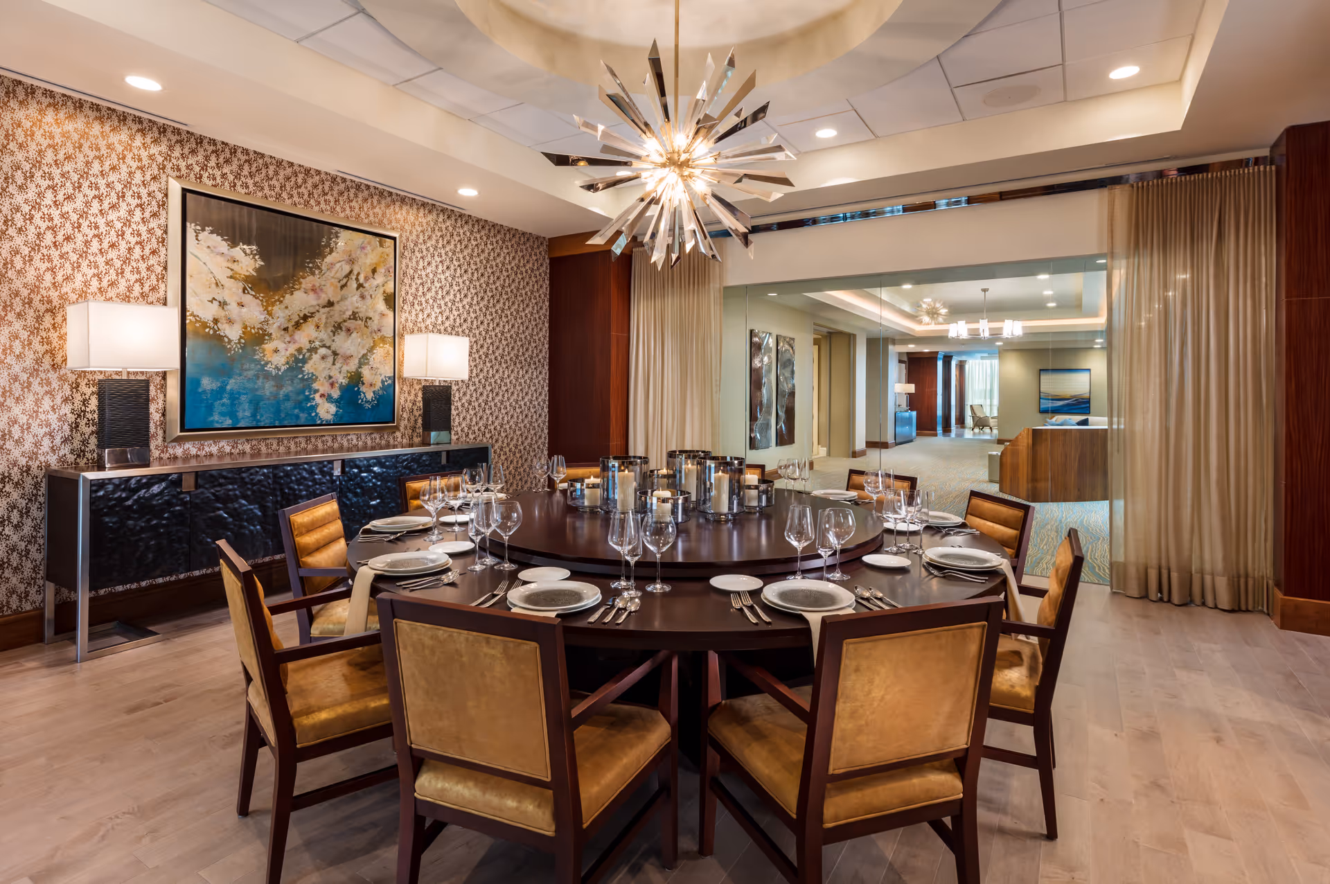 A luxurious dining room with a large round wooden table set with plates, glasses, and silverware. The table is surrounded by eight cushioned chairs with wooden frames. Above the table hangs a modern, starburst chandelier. The room features patterned wallpaper, a sideboard with two lamps, and a large floral painting. A large window or glass partition reveals a hallway and another seating area in the background.