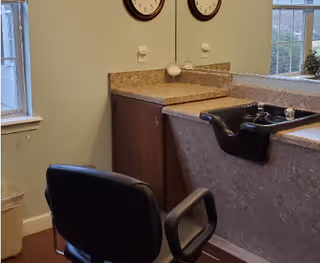 A salon-style hair-washing station with a black chair, basin sink, countertop and mirrored wall by a window.