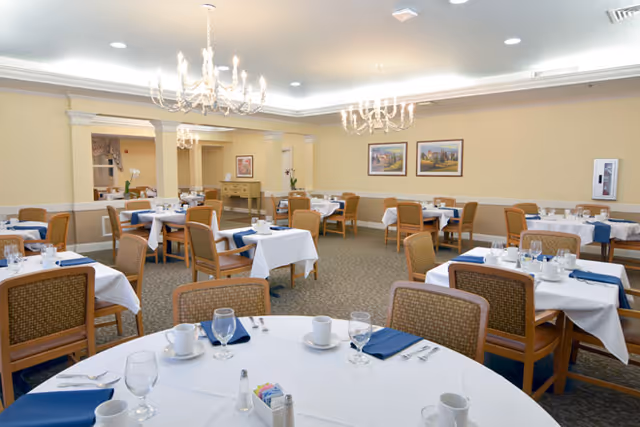 A spacious dining room with multiple tables covered in white tablecloths and set with cups, glasses, silverware, and blue napkins. The room features beige walls, carpeted floors, two chandeliers, framed artwork on the walls, and wooden chairs with woven backs.