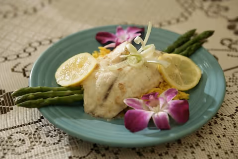 A plated meal featuring a cooked white fish fillet garnished with lemon slices, asparagus spears, and purple edible flowers, served on a blue plate placed on a lace tablecloth.