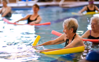 A group of elderly women participating in a water exercise class in an indoor swimming pool, each holding colorful pool noodles.