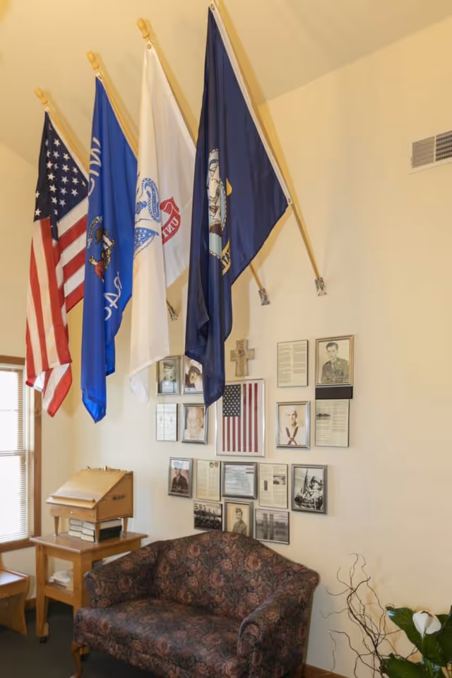 A corner of a room with four flags mounted on the wall, including the American flag and other military or organizational flags. Below the flags is a floral-patterned couch, and above it, a wall display of framed photographs, documents, and a small American flag. There is a wooden side table with a slanted top and some books underneath, next to a window with blinds. A plant with white flowers and some decorative branches is visible in the bottom right corner.
