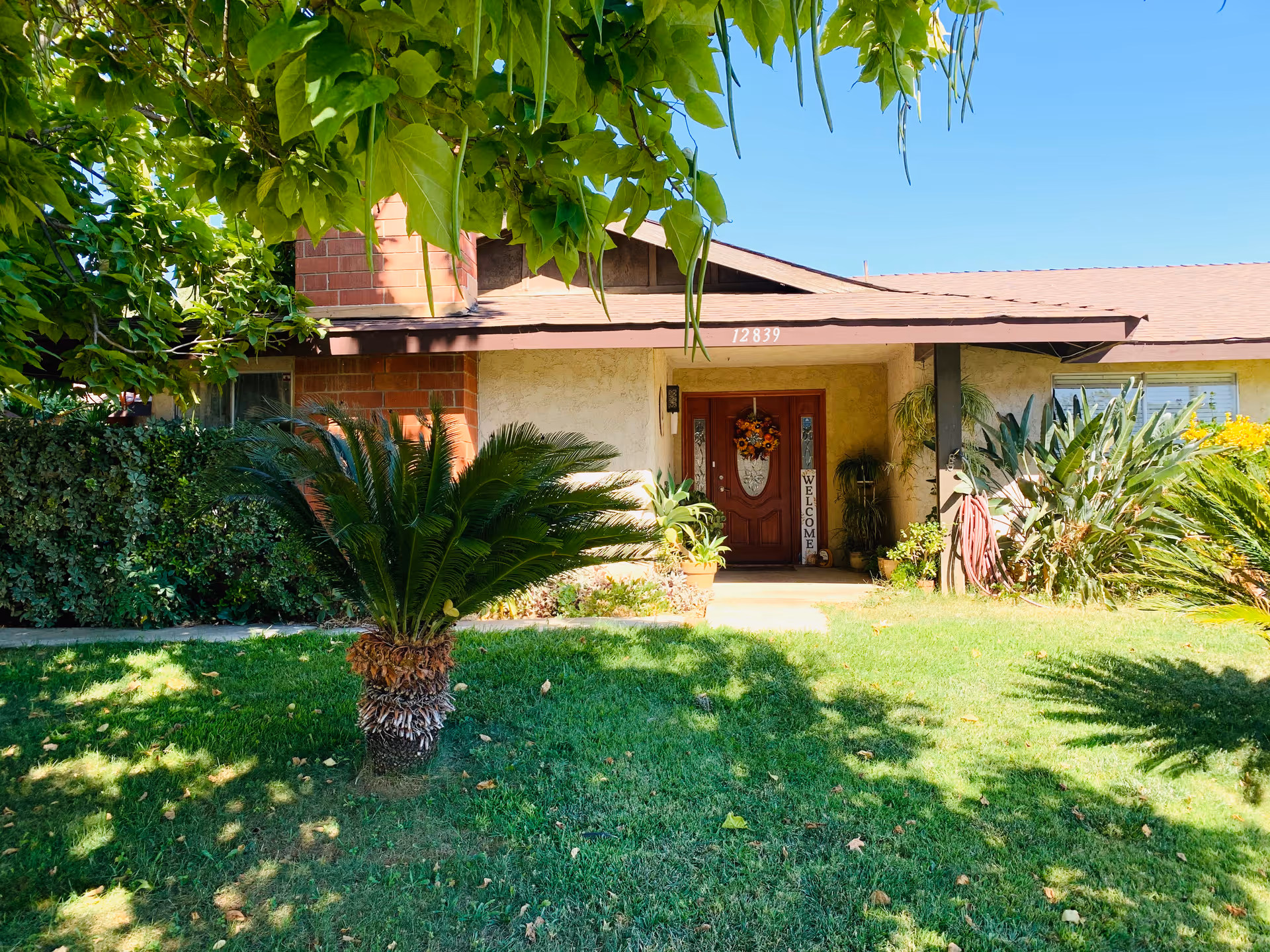 Front exterior of a single-story home with a covered porch, a wooden front door decorated with a wreath and a 'WELCOME' sign, and a green lawn with palm-like plants.