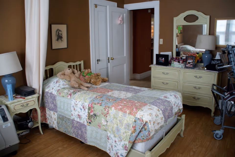A cozy bedroom with a single bed covered in a colorful patchwork quilt. Two teddy bears are placed on the bed. There is a nightstand with a blue lamp and some items on it next to the bed. A dresser with a mirror, lamp, and framed photos is against the wall. The room has wooden flooring and brown walls. A walker is visible near the dresser, and a door is open leading to another room.