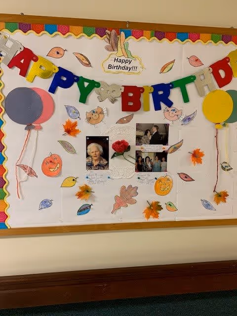A decorated bulletin board in a hallway with a "Happy Birthday" banner, photos, paper leaves, pumpkins and balloon cutouts.