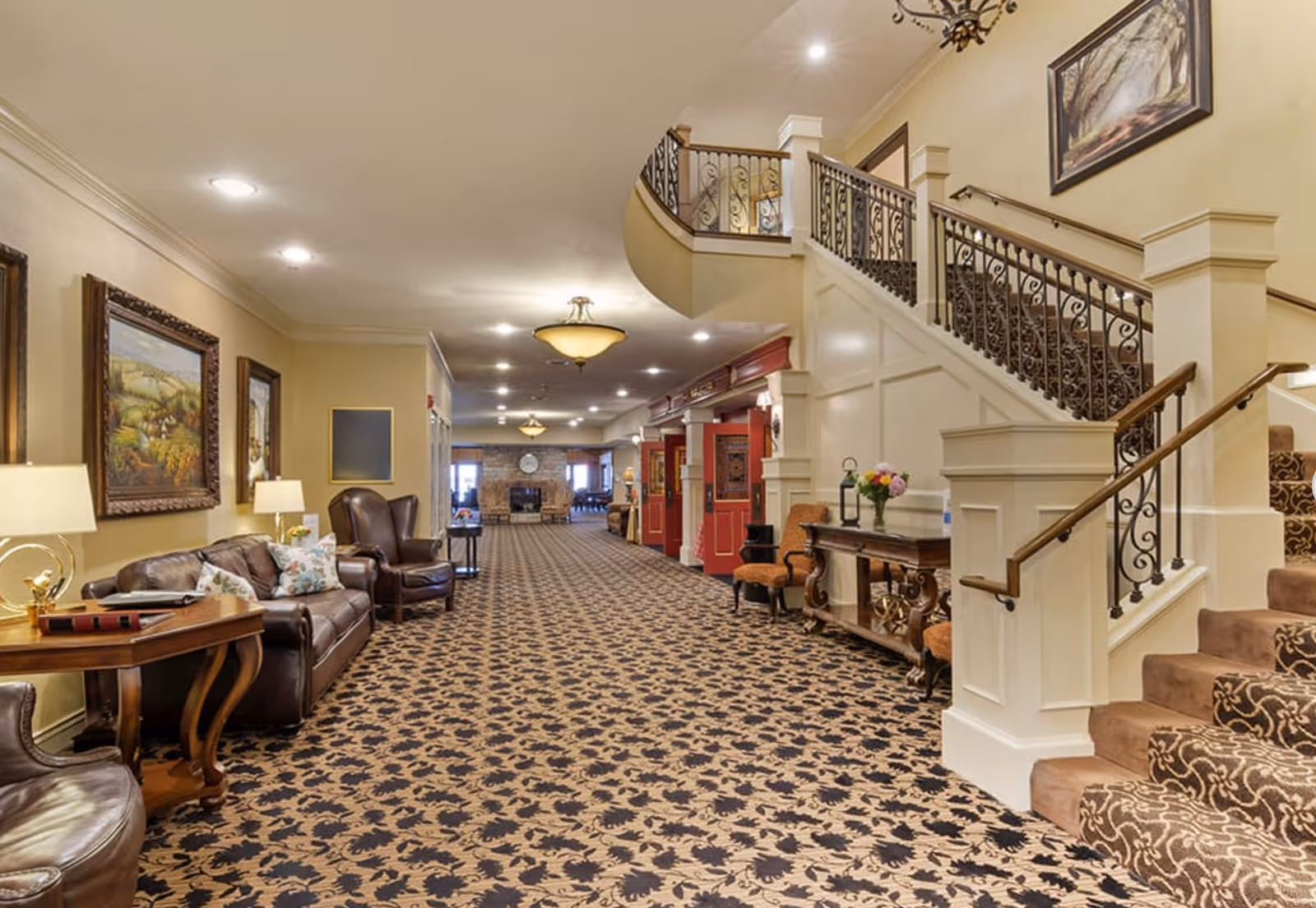 A spacious, well-lit interior hallway of a senior living facility featuring patterned carpet, leather sofas, armchairs, framed paintings on the walls, a staircase with ornate railings, and decorative tables with lamps and flowers.