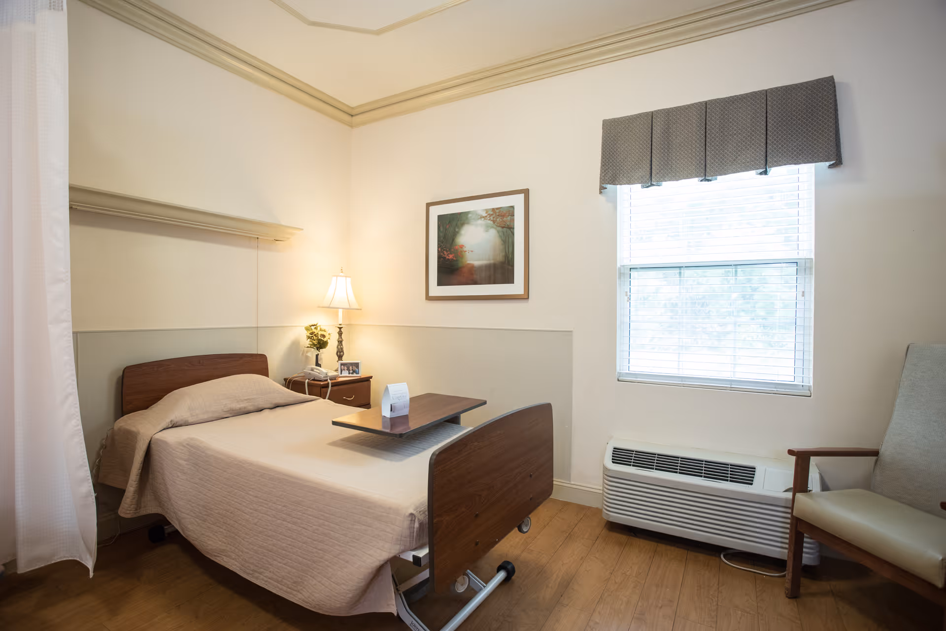 A small, neatly arranged bedroom in an assisted living facility featuring a single bed with beige bedding, a wooden bedside table with a lamp and a small plant, a framed picture on the wall, a window with blinds and a valance, a beige armchair, and a heating/cooling unit below the window.