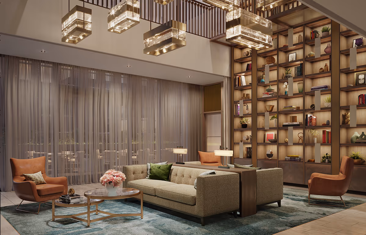 A modern and elegant living room area with a large beige sofa adorned with green and gray pillows, two brown leather armchairs, a round glass coffee table with a vase of pink flowers, and a tall wooden bookshelf filled with decorative items and books. The room features large sheer curtains, a blue patterned rug, and contemporary rectangular pendant lights hanging from the ceiling.