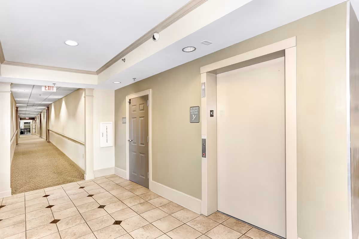 Interior hallway of a senior living facility with beige walls, tiled floor near an elevator, and carpeted floor extending down the corridor. The elevator door is closed, and there is a gray door next to it. The hallway is well-lit with ceiling lights and has handrails along the walls.