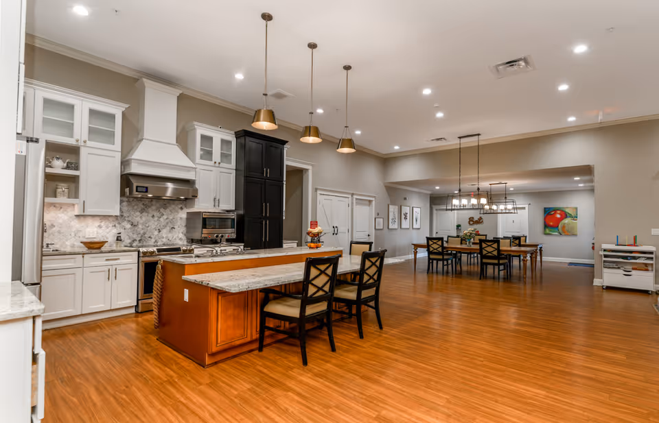 Spacious communal kitchen and dining area with a large island, pendant lights, and multiple dining tables on wood flooring.