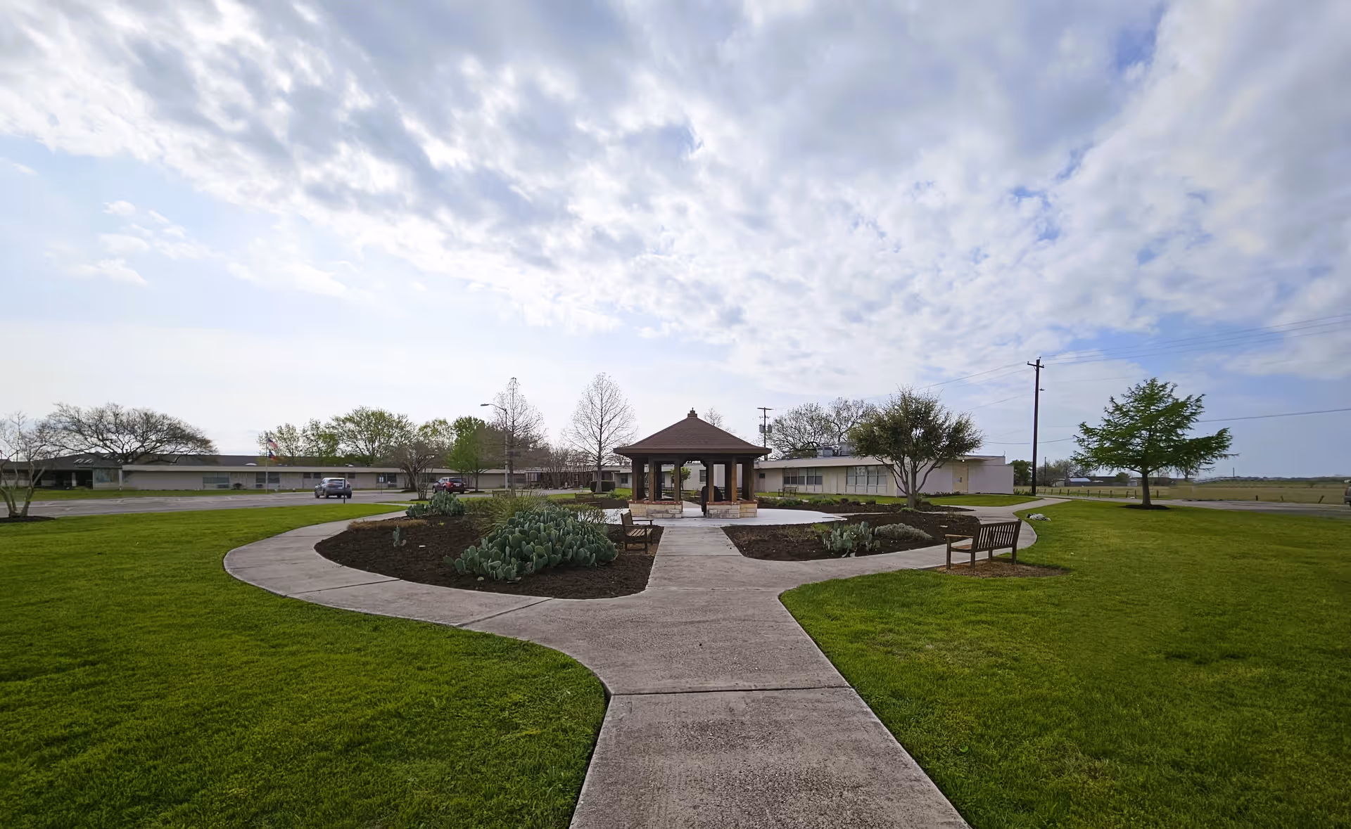 A wide outdoor garden area with a concrete pathway leading to a wooden gazebo in the center. The garden is surrounded by green grass, small trees, and benches. In the background, there is a single-story building under a partly cloudy sky.