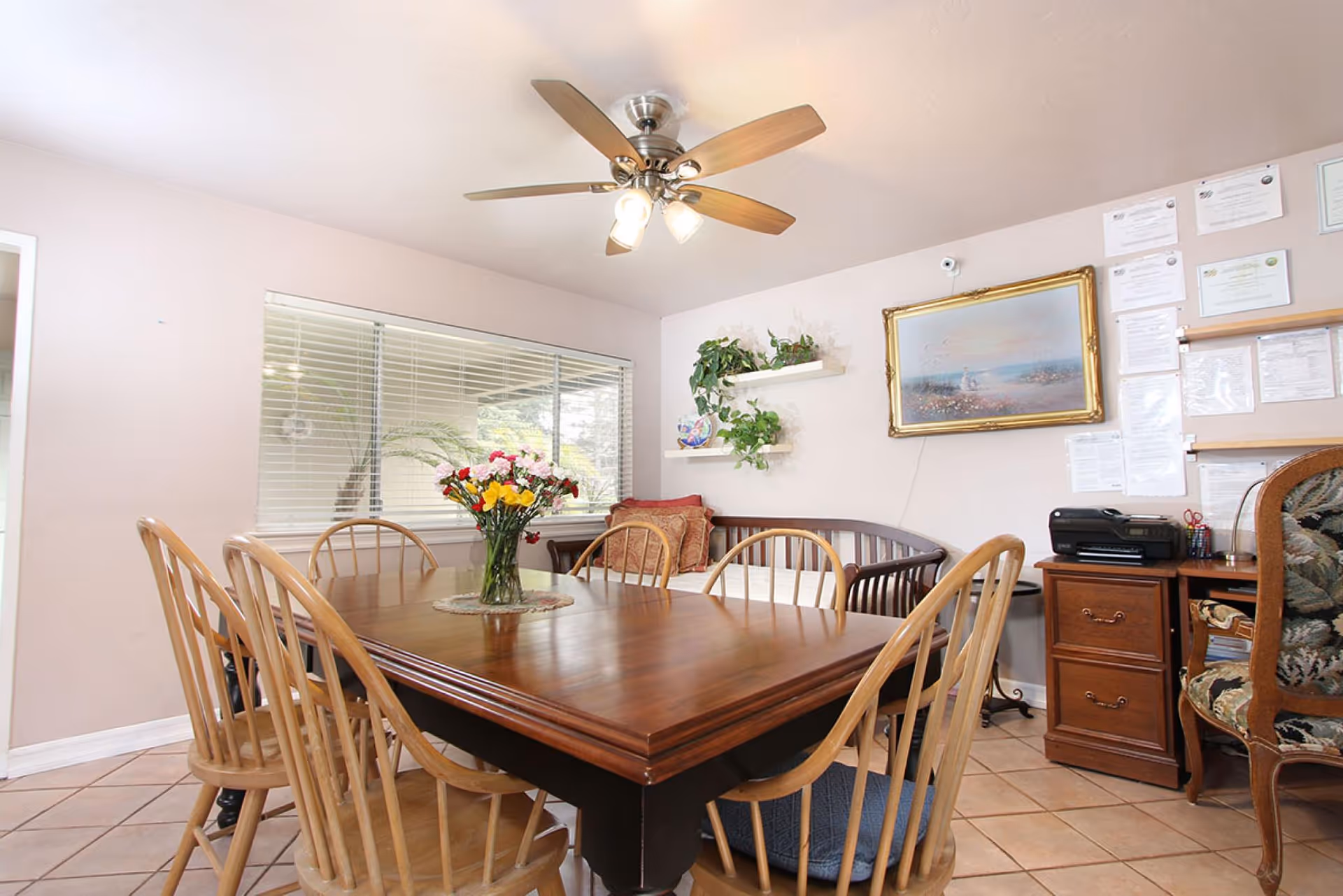 A dining room with a wooden table surrounded by six wooden chairs. A vase with colorful flowers is placed in the center of the table. The room has a ceiling fan with lights, a large window with blinds, and a corner with a wooden bench and cushions. There are two white shelves with green plants and decorative items on the wall, along with a framed painting and several documents pinned on a bulletin board. A wooden cabinet with a printer and office supplies is also visible.