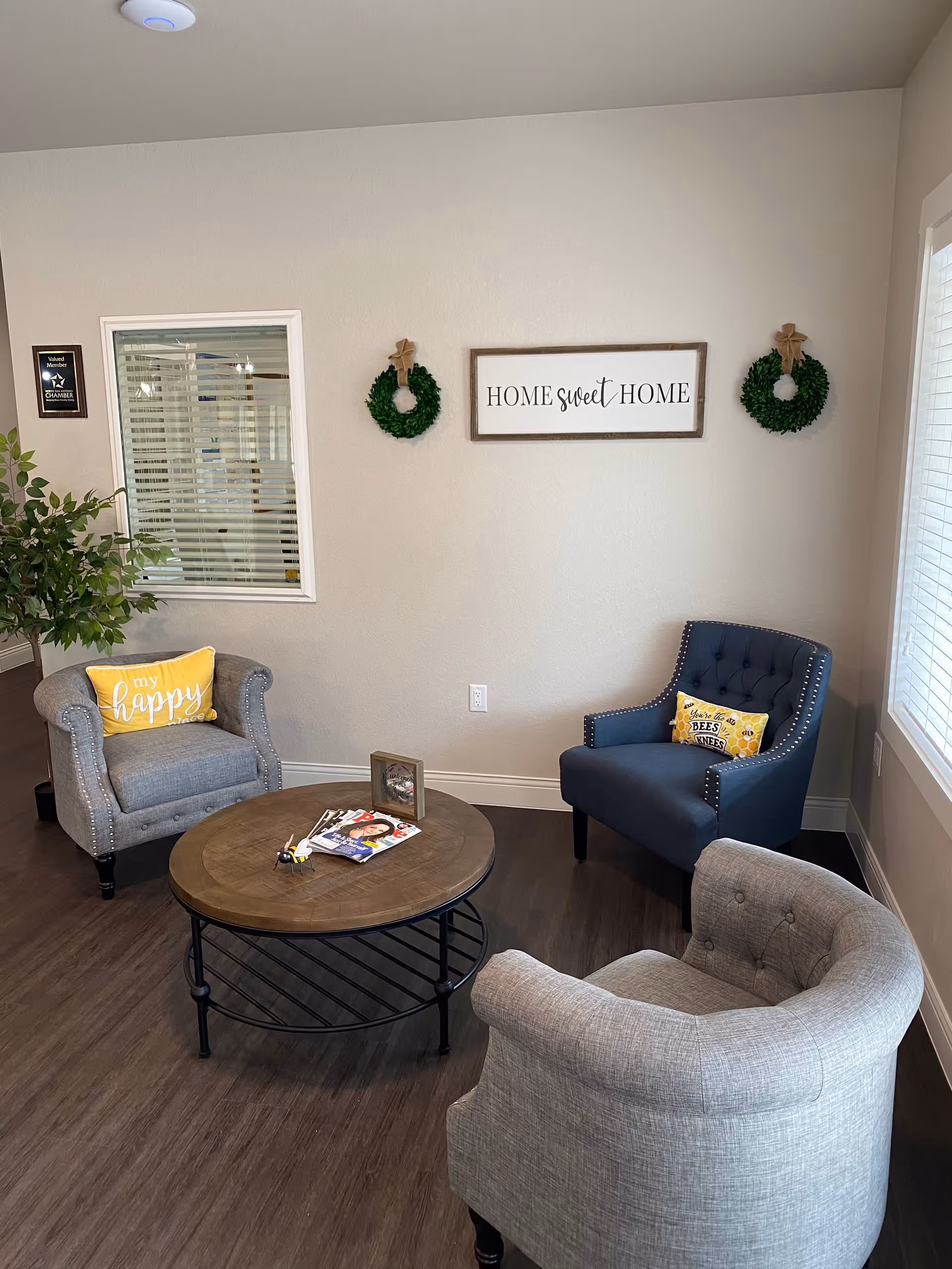 A cozy seating area in an assisted living facility with three upholstered armchairs arranged around a round wooden coffee table. The chairs have decorative pillows, one yellow with the words 'my happy place' and another with a bee theme. On the wall above the chairs is a sign that reads 'HOME sweet HOME' flanked by two small green wreaths. There is a window with blinds on the right and a small plant on the left.