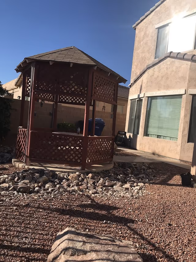 Outdoor area with a wooden gazebo surrounded by rocks and gravel, adjacent to a beige stucco building with windows and a clear blue sky overhead.