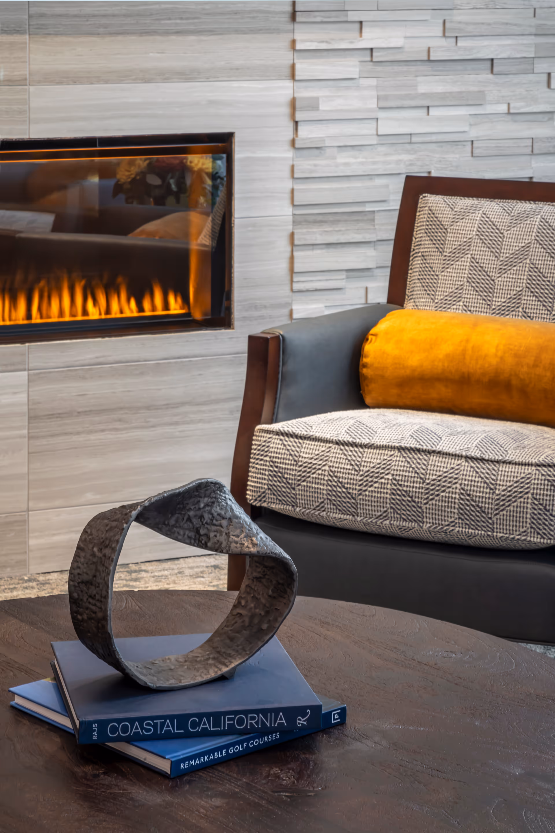 A cozy interior scene featuring a modern fireplace with flames visible, a patterned armchair with a mustard yellow cylindrical pillow, and a dark wooden coffee table with two books titled 'Coastal California' and 'Remarkable Golf Courses' stacked on it. On top of the books is a decorative metal sculpture. The wall behind the fireplace has a textured, light-colored tile design.