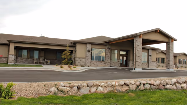 Front exterior of a single-story senior living facility with a covered entrance, stone facade, and surrounding landscaping.