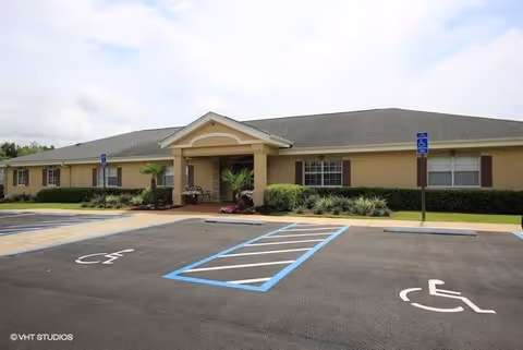 Single-story beige building with a gray roof and a covered entrance, surrounded by greenery and a parking lot with designated handicapped parking spaces in front.
