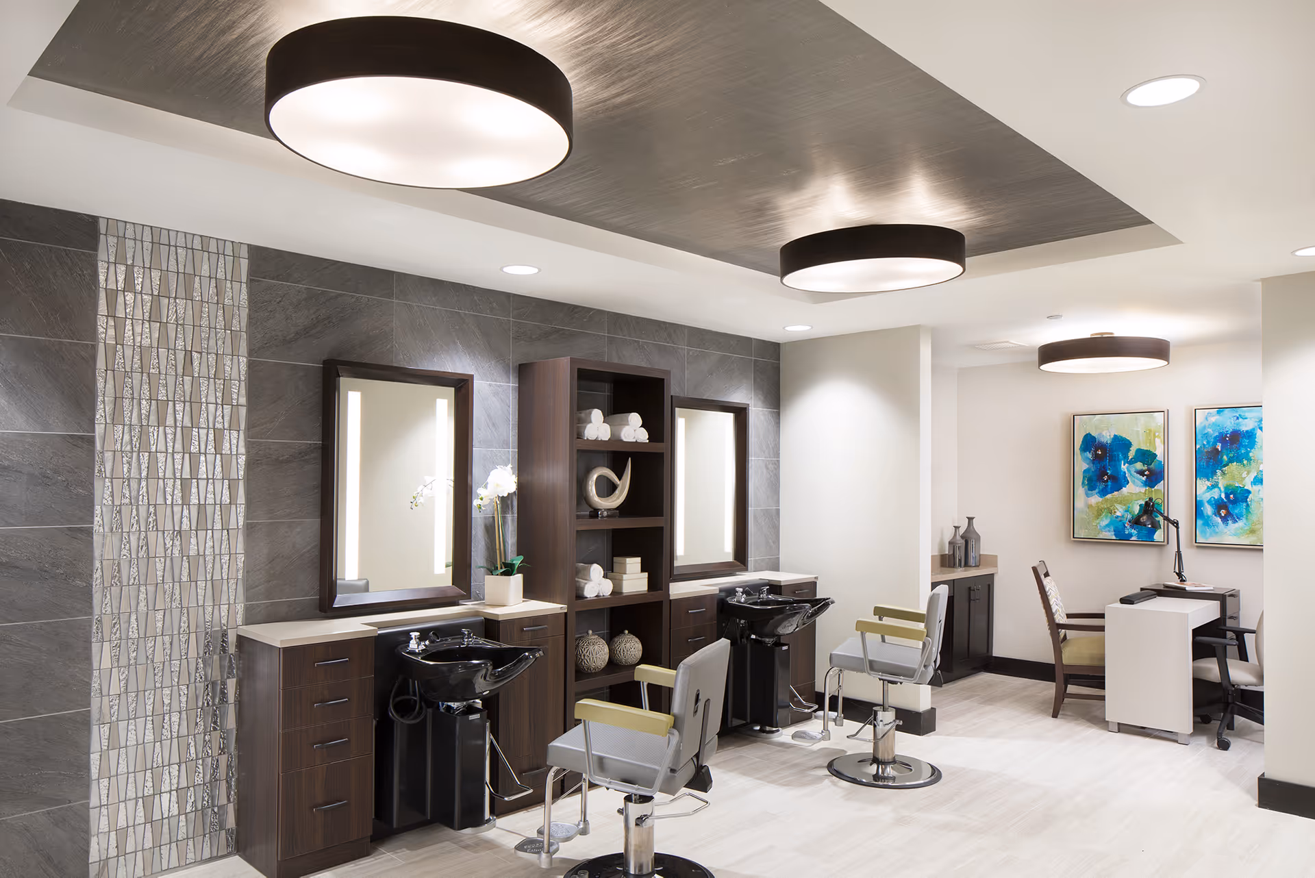 Modern salon area with two styling stations featuring large mirrors, black wash basins, and salon chairs. The space has a sleek design with dark wood cabinetry, decorative tile accents on the wall, and contemporary ceiling lights. In the background, there is a small seating area with a desk, chairs, and colorful abstract artwork on the wall.