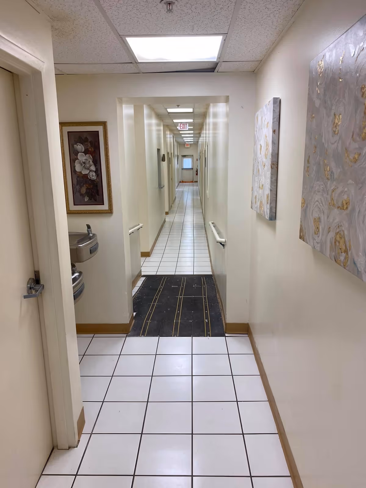 A long, narrow hallway in a personal care home with white tiled floors and cream-colored walls. There are handrails on both sides of the hallway, floral paintings on the walls, and a water fountain on the left side near the entrance. The ceiling has fluorescent lighting panels, and an exit sign is visible at the far end of the corridor.