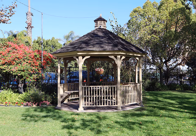 A wooden gazebo with a shingled roof situated on a well-maintained grassy lawn, surrounded by trees, flowering bushes, and a black metal fence in the background under a clear blue sky.