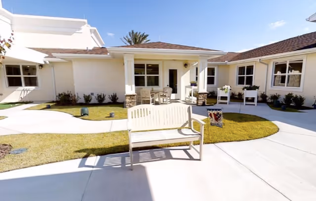 Outdoor courtyard area of a senior living facility with a white bench in the foreground, concrete walkways, green grass patches, and a covered patio with chairs and a table. The building is single-story with white walls and multiple windows under a clear blue sky.