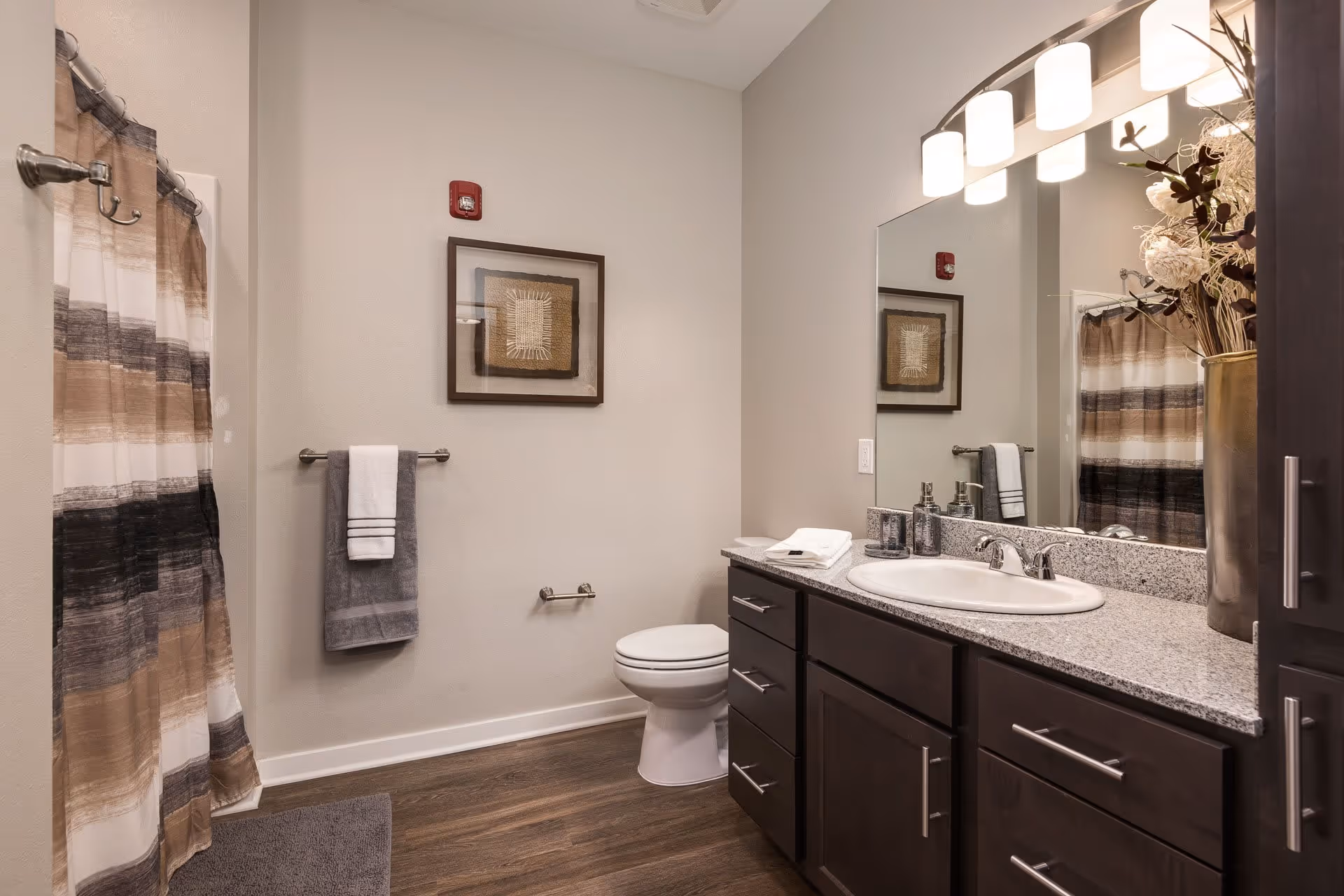 A modern bathroom featuring a shower with a striped brown, beige, and black curtain, a white toilet, a dark wood vanity with a granite countertop, a white sink, and a large mirror above. The wall has a framed decorative artwork and a towel rack with gray and white towels. The floor is wood-style, and there is a vase with dried flowers on the countertop.