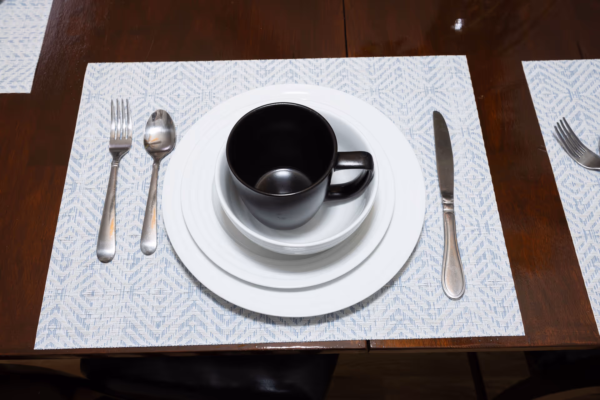 A dining table set with a white patterned placemat, a stack of three white plates of different sizes, and a black coffee mug on top. To the left of the plates are a fork and a spoon, and to the right is a knife. The table is wooden and dark brown.
