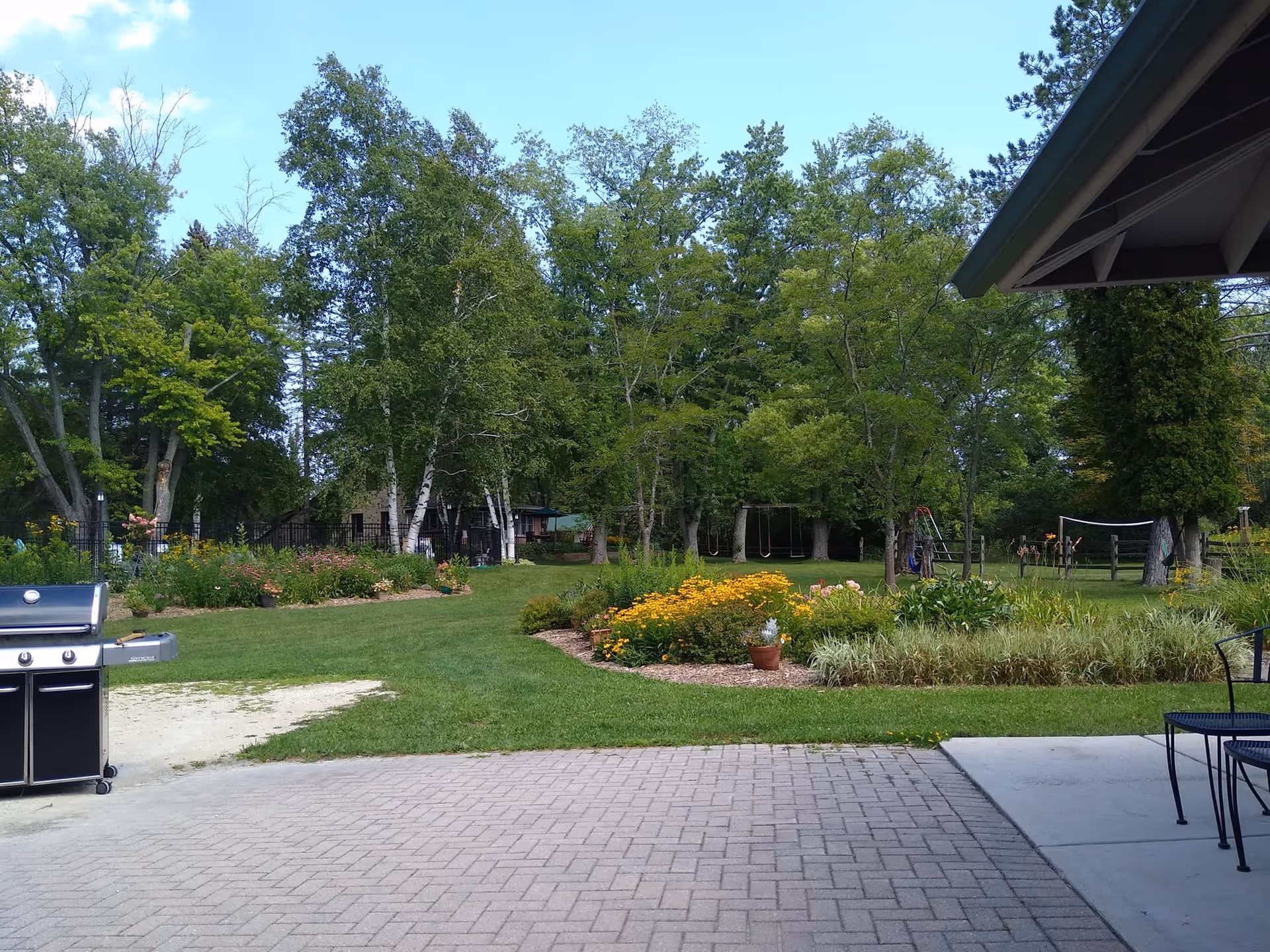 View of a garden area with green grass, colorful flower beds, and tall trees under a blue sky. There is a black grill on the left side and a patio area with metal chairs on the right side, partially covered by a roof.