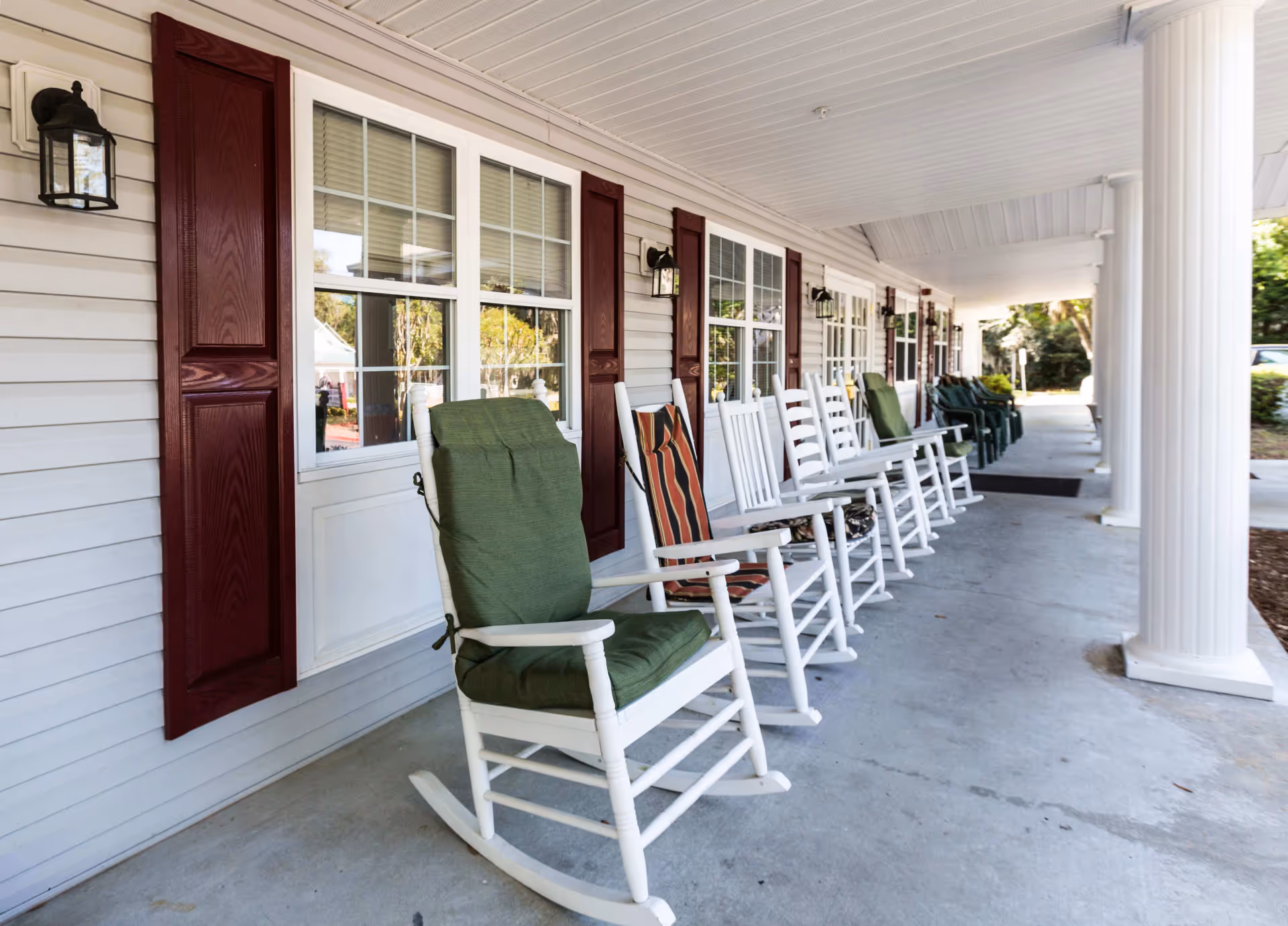 A covered porch area with a row of white rocking chairs, some with cushions, lined up against a building with white siding and red shutters. The porch has white columns and outdoor wall lanterns mounted beside the windows.